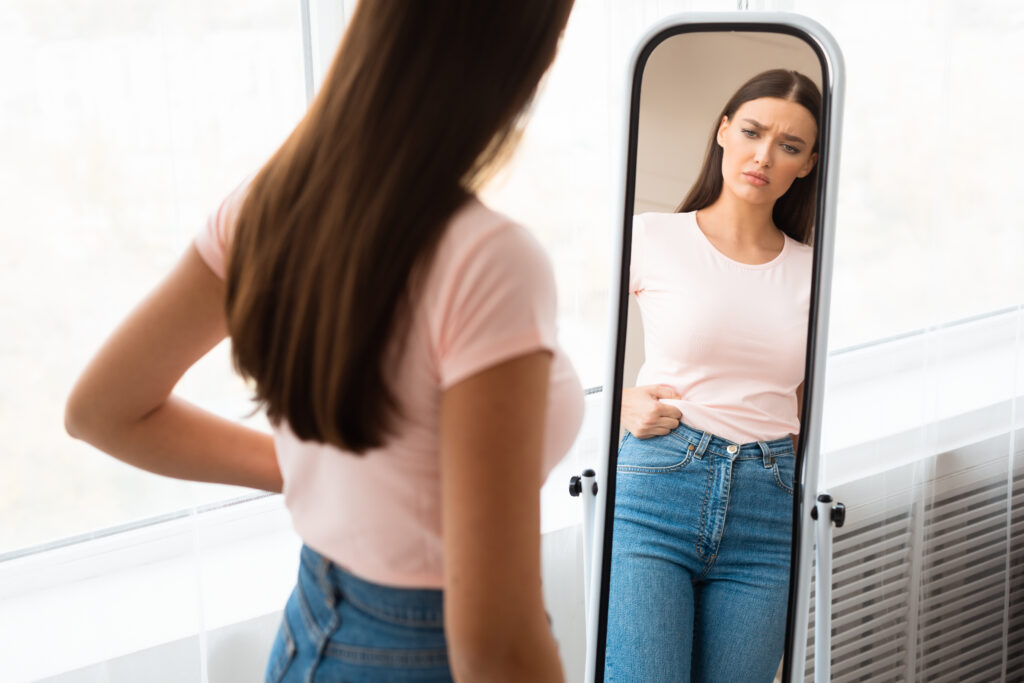 Salt Lake City woman pinches her belly fat, frowning deeply in the full-length mirror as she wonders about hormone imbalance and weight gain